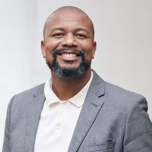 Smiling man in a gray suit and white shirt, standing against a plain background.